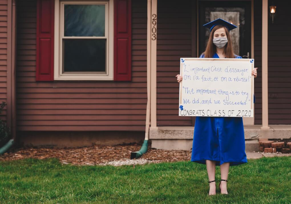 A student stands in her front lawn wearing a graduation cap and gown and face mask. She holds a sign with a handwritten message on congradulations 